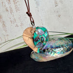 Herzförmiger Anhänger aus Olivenholz mit aquamarinfarbenem Harz und eingelegten Paua-Muschelstücken (Abalone), irisierender Blau-Grün-Schimmer. Der Anhänger hängt an einem braunen, verstellbaren Lederband mit Schiebeknoten; Aufhängung aus Edelstahl. Produktfoto vor maritimer Deko/Abalone-Schale.
