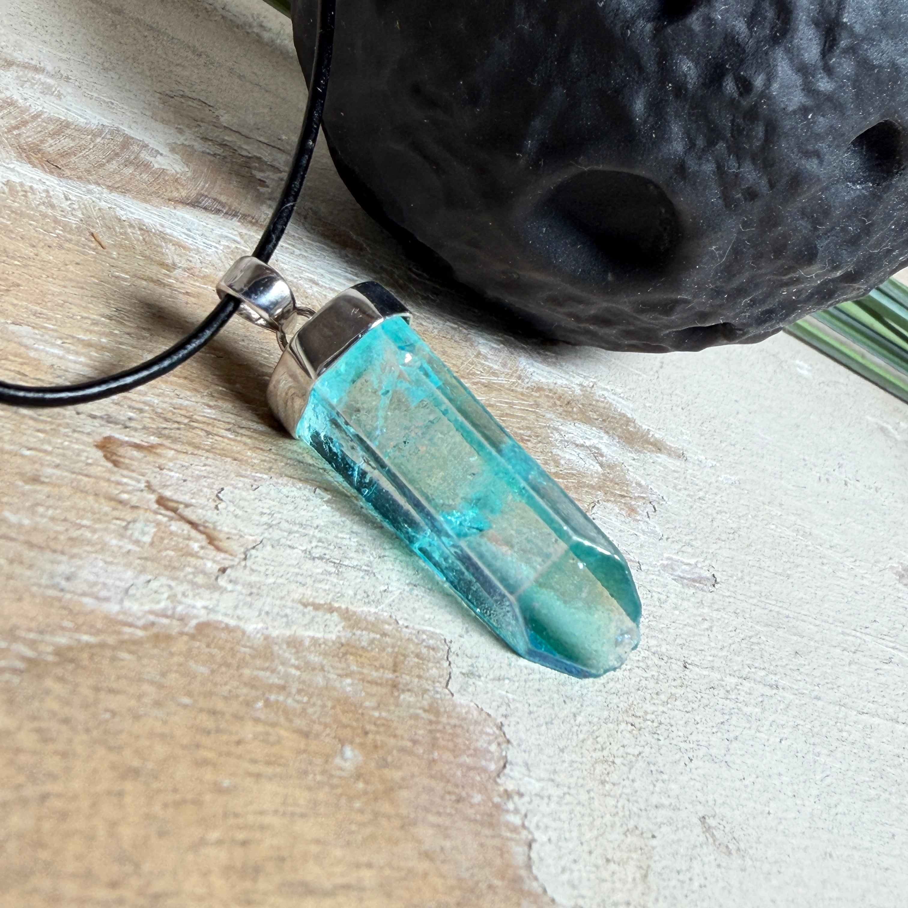 Nahaufnahme einer handgefertigten Halskette mit Aqua Aura Rohkristall: naturbelassener, sechskantiger Bergkristall mit goldbedampfter Aqua-Beschichtung in Türkisblau, gefasst in 925er Silber und getragen an einer schwarzen Lederkette mit Edelstahl-Karabiner und Verlängerungskettchen.