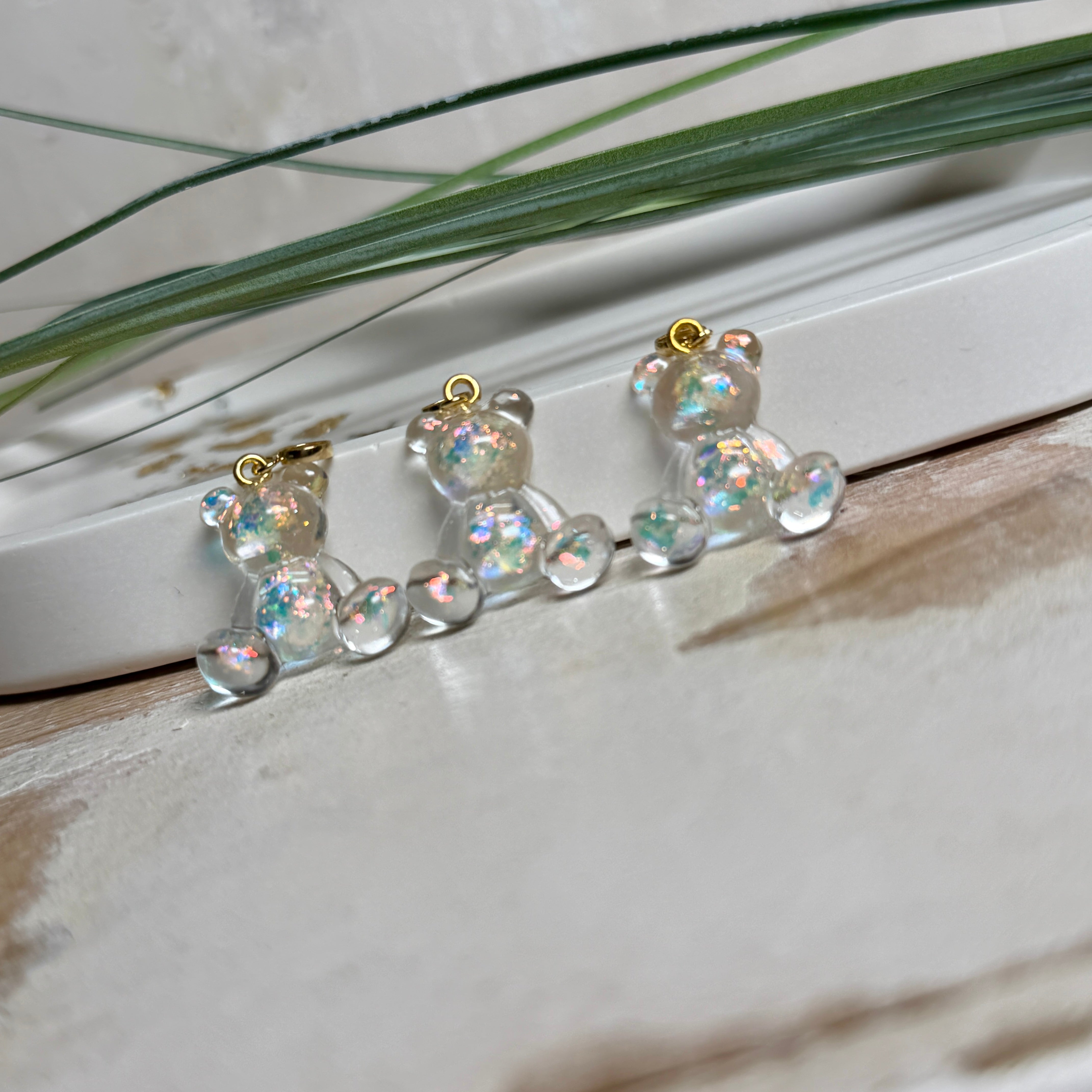 Kleiner Bärchen-Anhänger aus transparentem Harz mit irisierenden Flakes in Regenbogen-Schimmer, goldfarbene Anhängerschlaufe aus Edelstahl. Größe ca. 2,2 × 2,1 cm. Handgefertigtes Unikat; Flakes-Anordnung variiert, vereinzelt kleine Luftbläschen möglich.