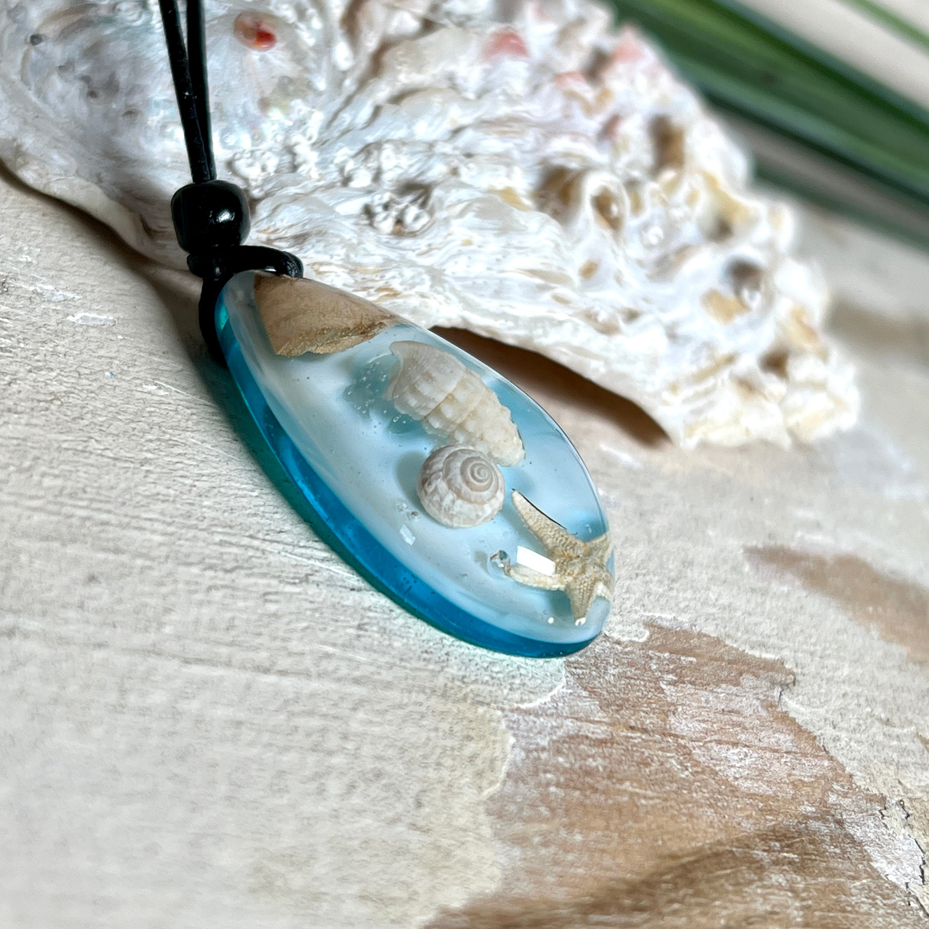 Handgefertigter Tropfenanhänger aus Maserholz und klarem Epoxidharz in Aquatönen. Im Harz sind echte Muscheln und ein kleiner Seestern eingebettet. Durch die besondere Mehrschicht-Gießtechnik wirkt es, als würden die Muscheln im Wasser schweben. Größe ca. 3,8 × 2 cm. Verstellbares Lederband (24–44 cm) mit Holzperlen. Maritimes Unikat.