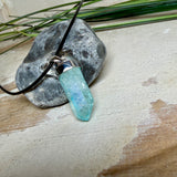 Nahaufnahme einer handgefertigten Halskette mit Aqua Aura Rohkristall: naturbelassener, sechskantiger Bergkristall mit goldbedampfter Aqua-Beschichtung in Türkisblau, gefasst in 925er Silber und getragen an einer schwarzen Lederkette mit Edelstahl-Karabiner und Verlängerungskettchen.