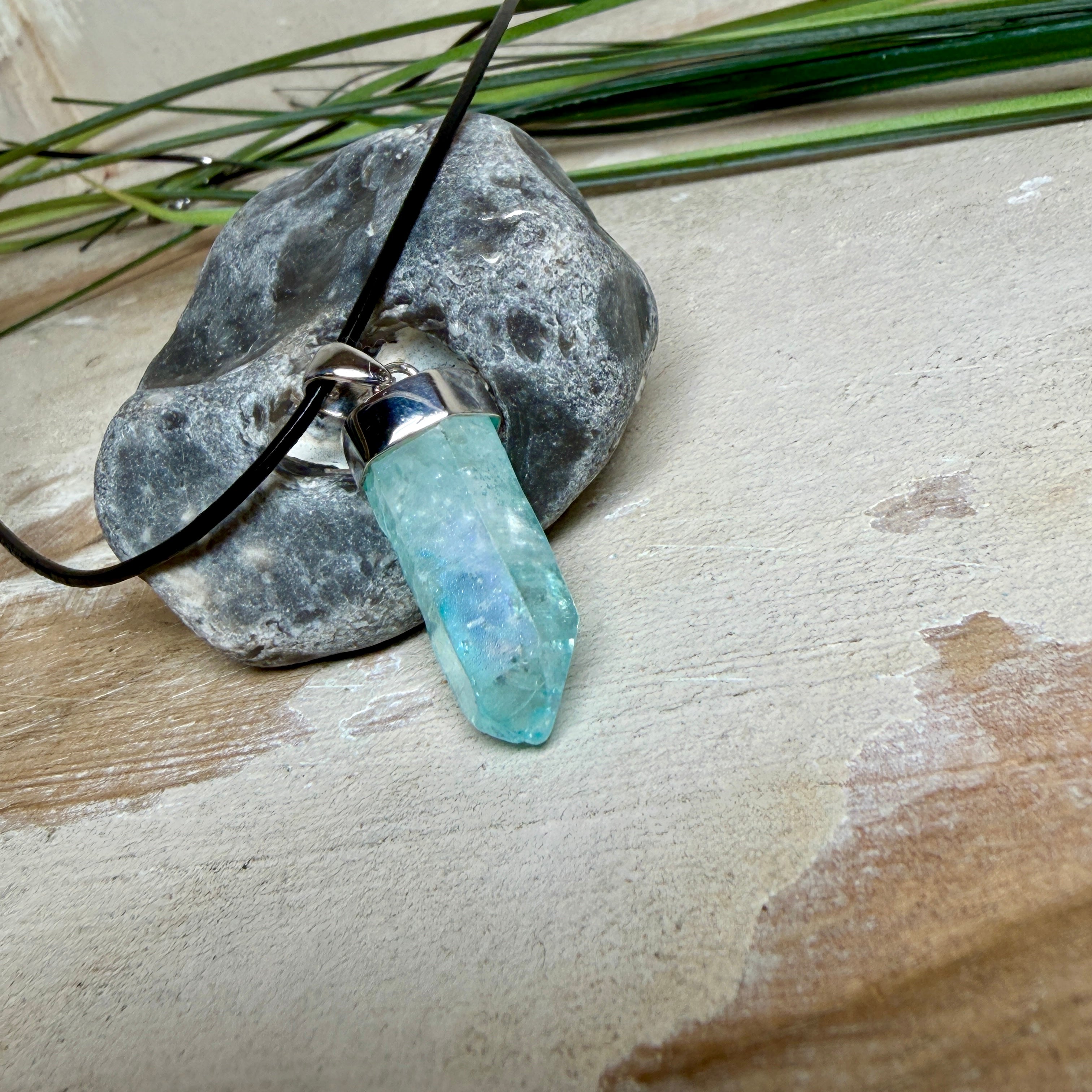 Nahaufnahme einer handgefertigten Halskette mit Aqua Aura Rohkristall: naturbelassener, sechskantiger Bergkristall mit goldbedampfter Aqua-Beschichtung in Türkisblau, gefasst in 925er Silber und getragen an einer schwarzen Lederkette mit Edelstahl-Karabiner und Verlängerungskettchen.