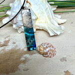Rechteckiger Stabanhänger aus Maserholz und glasklarem Epoxidharz in tiefen Blau- und Türkistönen mit organischer Lagunen/Riff-Optik. Hochglanzpoliert, ca. 5,3 × 1,2 cm. Der Anhänger hängt an einer schwarzen Lederkette mit Edelstahl-Karabiner und 5-cm-Verlängerung (45 + 5 cm); die Anhängerschlaufe ist ebenfalls aus Edelstahl. Handgefertigtes Unikat, Lieferung in edler Schmuckbox – maritimer Look.