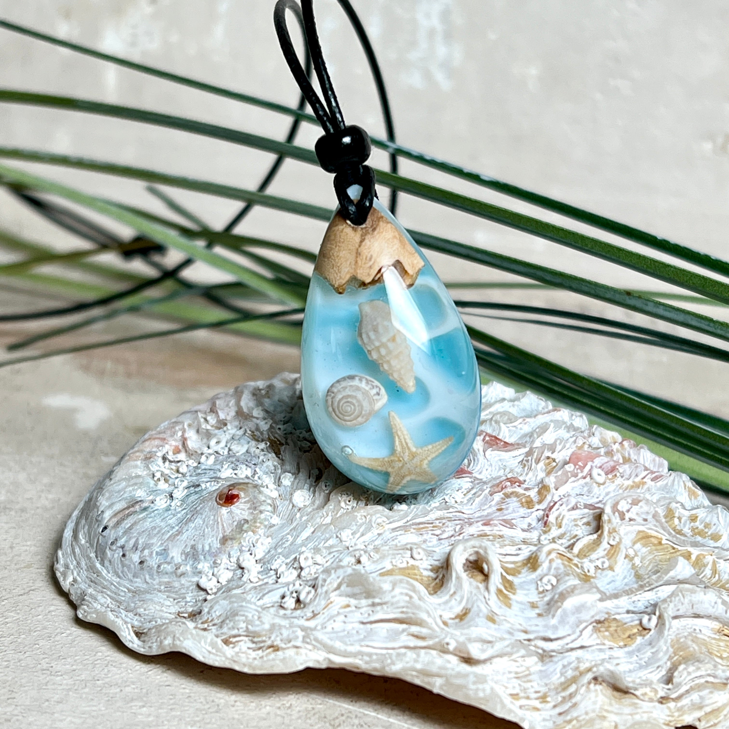 Handgefertigter Tropfenanhänger aus Maserholz und klarem Epoxidharz in Aquatönen. Im Harz sind echte Muscheln und ein kleiner Seestern eingebettet. Durch die besondere Mehrschicht-Gießtechnik wirkt es, als würden die Muscheln im Wasser schweben. Größe ca. 3,8 × 2 cm. Verstellbares Lederband (24–44 cm) mit Holzperlen. Maritimes Unikat.