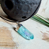 Nahaufnahme einer handgefertigten Halskette mit Aqua Aura Rohkristall: naturbelassener, sechskantiger Bergkristall mit goldbedampfter Aqua-Beschichtung in Türkisblau, gefasst in 925er Silber und getragen an einer schwarzen Lederkette mit Edelstahl-Karabiner und Verlängerungskettchen.