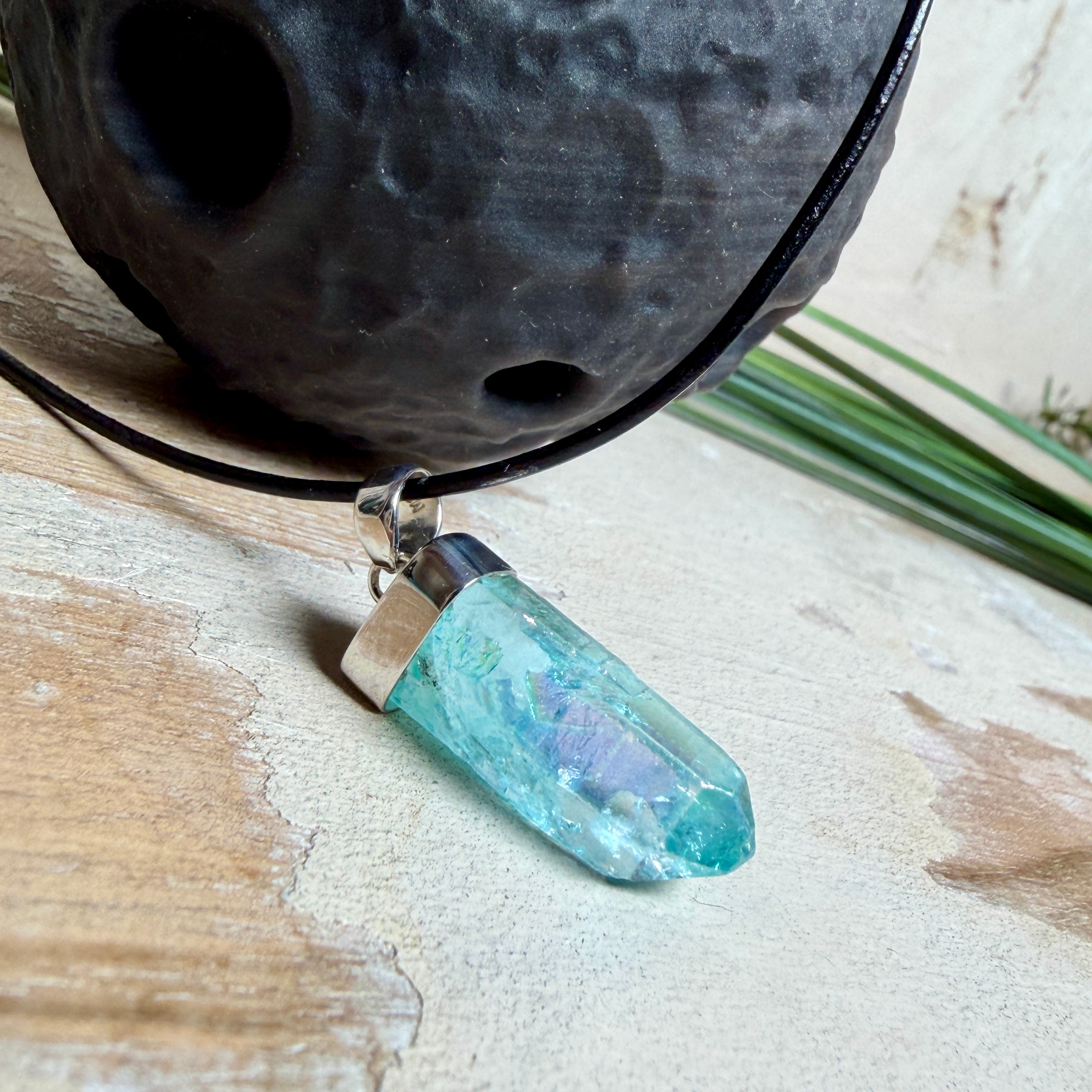Nahaufnahme einer handgefertigten Halskette mit Aqua Aura Rohkristall: naturbelassener, sechskantiger Bergkristall mit goldbedampfter Aqua-Beschichtung in Türkisblau, gefasst in 925er Silber und getragen an einer schwarzen Lederkette mit Edelstahl-Karabiner und Verlängerungskettchen.
