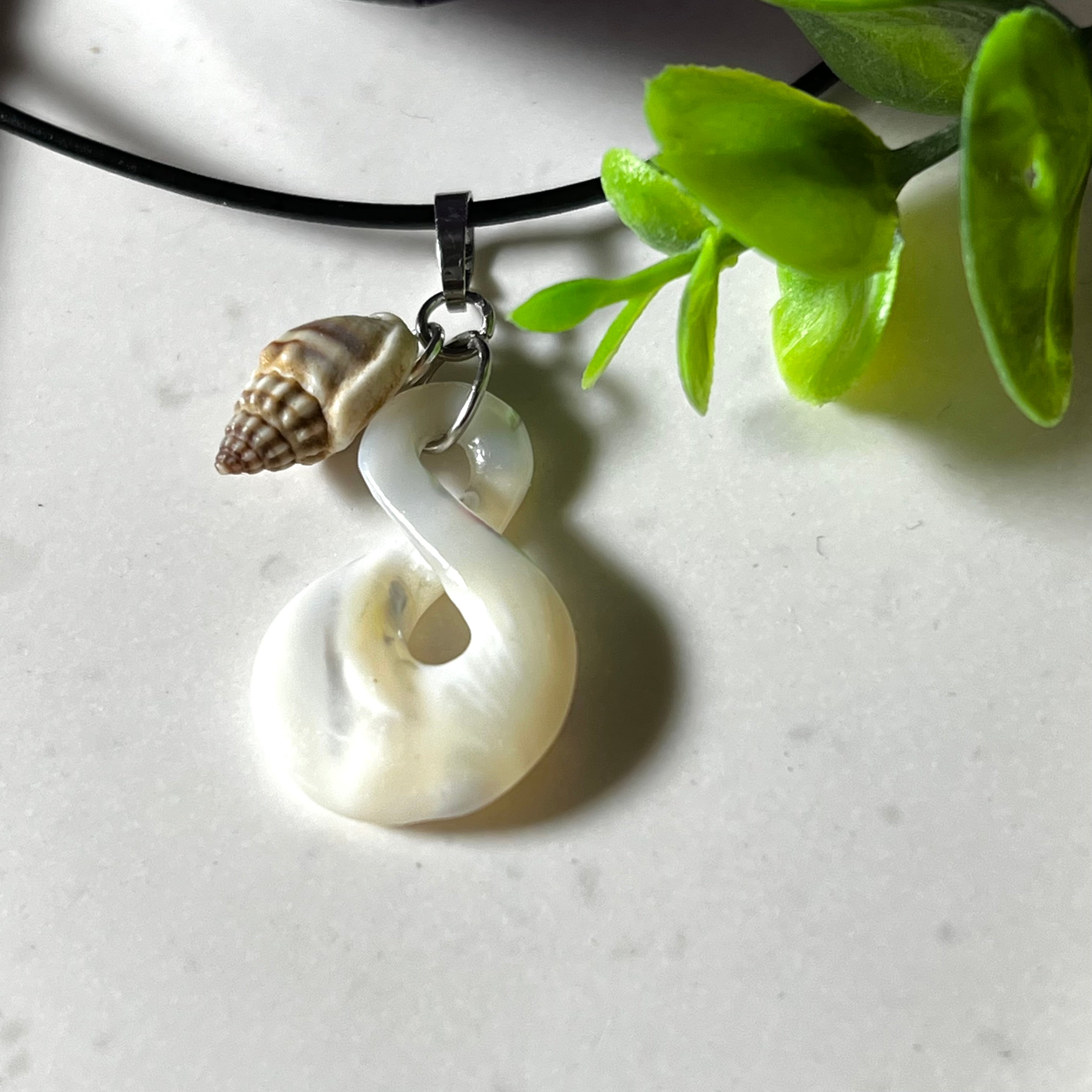 Detailaufnahme eines handgefertigten Anhängers aus echtem Perlmutt in Schlaufenform, kombiniert mit einer kleinen echten Muschel als Charm-Anhänger. Der Schmuck hängt an einer feinen schwarzen Lederschnur mit Edelstahlverschluss und symbolisiert Harmonie und Unendlichkeit. Ideal für Meeresliebhaberinnen und Fans von Naturmaterialien.