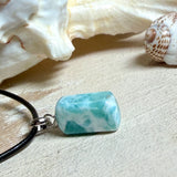 Larimar Freeform Anhänger an schwarzer Lederkette. Natürlicher, rechteckiger Larimar-Stein (ca. 1,5 × 1 cm) in Türkis-Blau mit weißen Strukturen, eingefasst in eine rhodinierte Silberöse. Beidseitig tragbar, an 45 cm Lederkette mit Edelstahl-Karabinerverschluss und 5 cm Verlängerung. Maritimes, zurückhaltend elegantes Schmuckstück mit der Optik karibischen Meerwassers.
