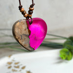 Handgefertigter Herzanhänger aus Olivenholz und leuchtend pinkem Epoxidharz mit eingeschlossenen Pusteblumensamen. Verstellbares braunes Lederband mit Holzperlen, Anhänger ca. 3,5 × 3,3 cm groß. Das Harz leuchtet dezent unter Schwarzlicht. Ein einzigartiges Unikat von ZeitlosSchmuckDesign, inspiriert von Kindheitsglück und Pusteblumen.