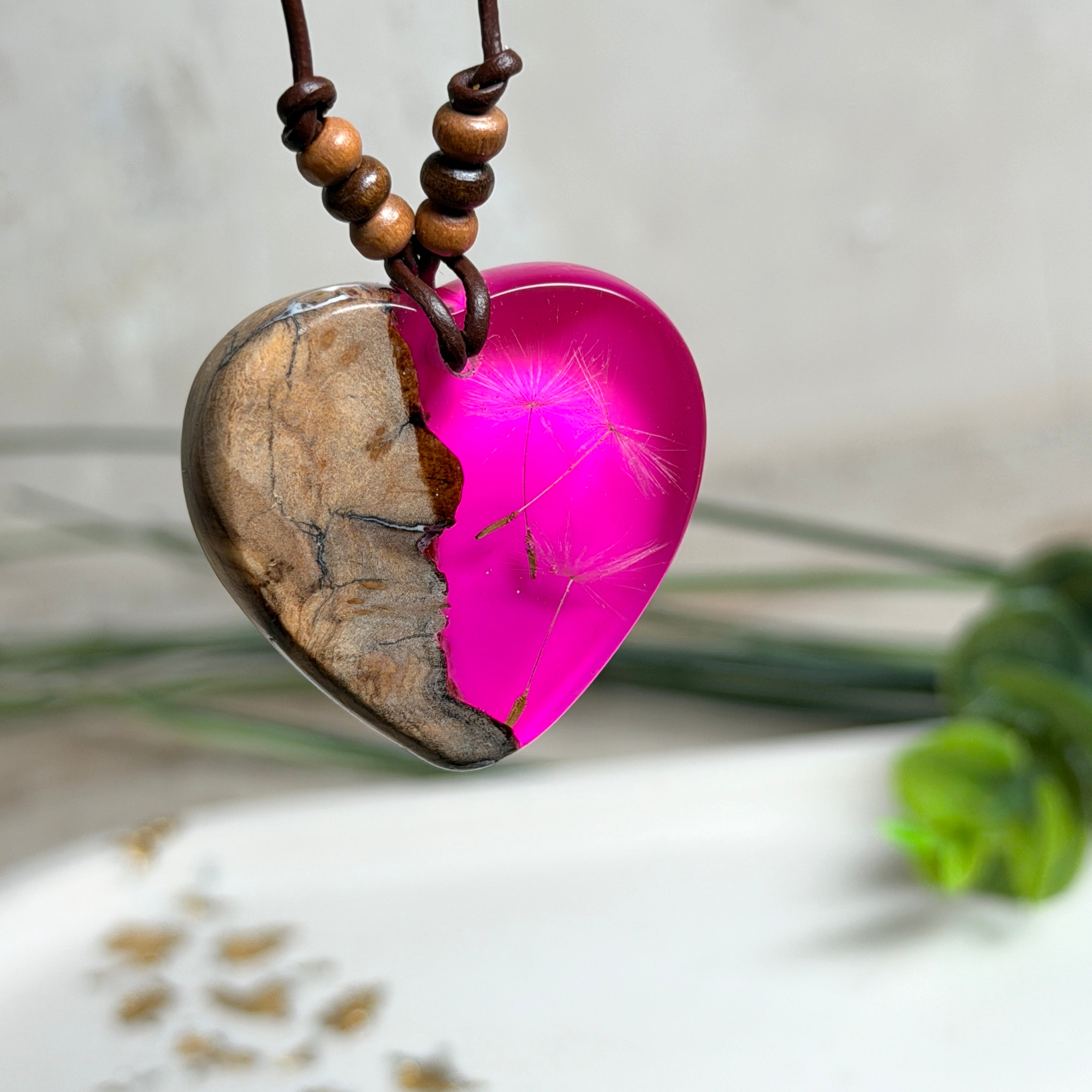 Handgefertigter Herzanhänger aus Olivenholz und leuchtend pinkem Epoxidharz mit eingeschlossenen Pusteblumensamen. Verstellbares braunes Lederband mit Holzperlen, Anhänger ca. 3,5 × 3,3 cm groß. Das Harz leuchtet dezent unter Schwarzlicht. Ein einzigartiges Unikat von ZeitlosSchmuckDesign, inspiriert von Kindheitsglück und Pusteblumen.