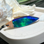 Beidseitig tragbarer Surfbrett-Anhänger aus Olivenholz, kombiniert mit klaren Epoxidharz in Blau-Grün. Eingearbeiteter synthetischer Opal erzeugt ein faszinierendes Farbspiel wie Nordlichter. Größe ca. 6 × 1,8 cm. Mit verstellbarem Lederband (26–45 cm), dekorativen Perlen & Schiebeknoten. Handgefertigt & einzigartig. Ein Schmuckstück mit Naturinspiration.