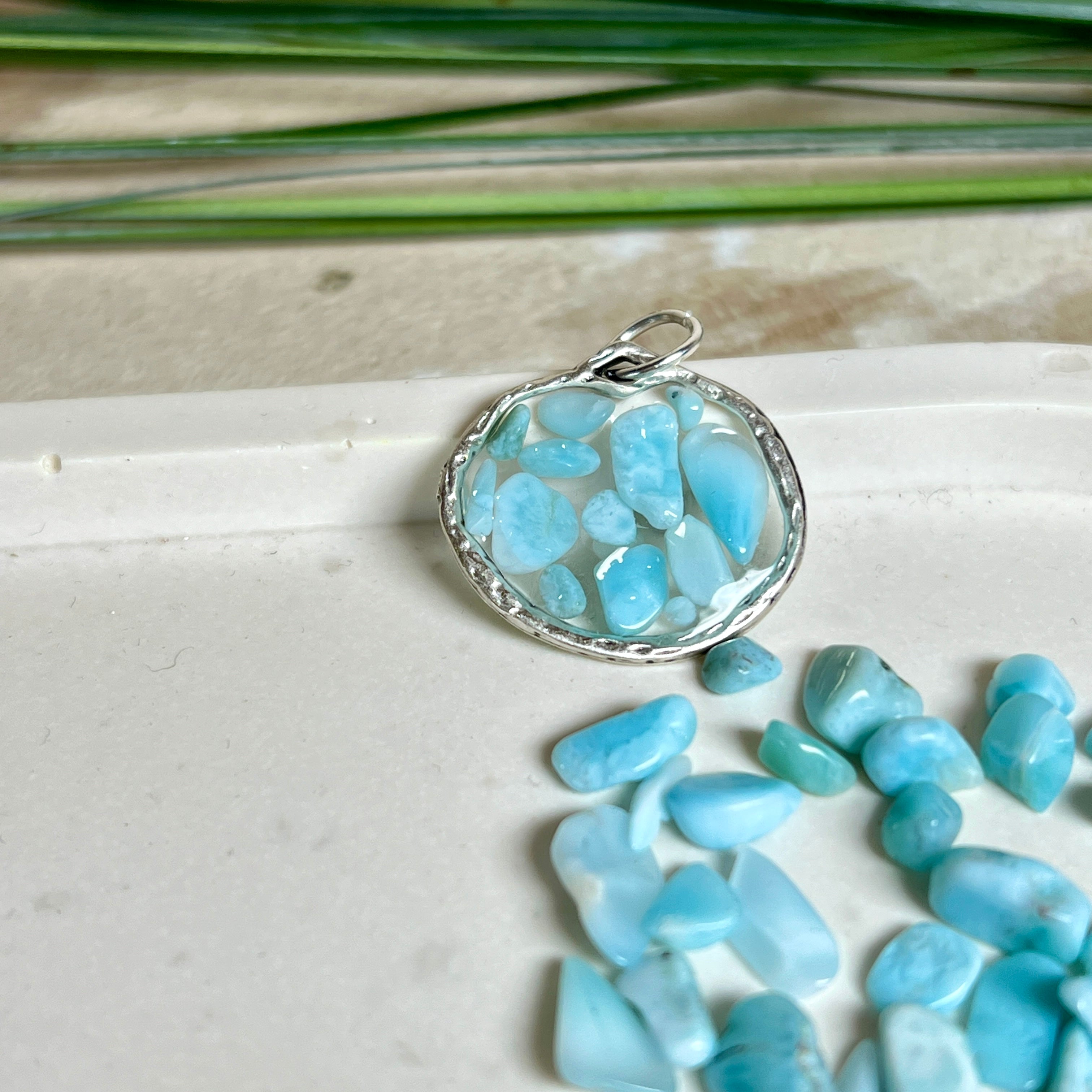 Runder Anhänger „Atlantis Dream“ aus antik-silberfarbener Zinklegierung, gefüllt mit kleinen Larimar-Edelsteinchen und klarem Resin. Die Larimarsteine zeigen sanfte Blau- und Weißtöne, die an das Meer erinnern. Durchmesser ca. 22 mm, handgefertigtes Unikat, fotografiert auf maritimem Untergrund mit kleinen Larimarsteinen und Muscheln zur Größenorientierung.