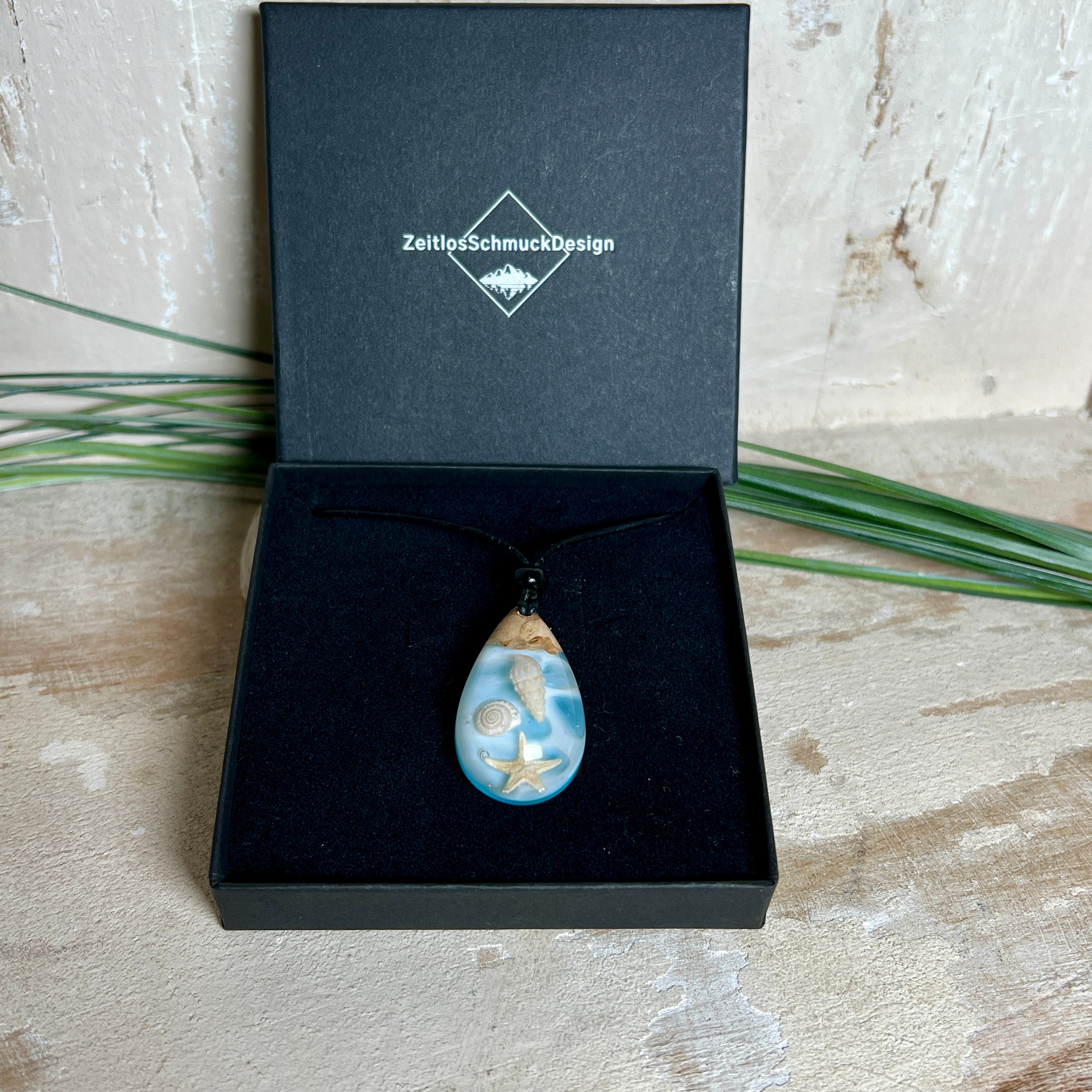 Handgefertigter Tropfenanhänger aus Maserholz und klarem Epoxidharz in Aquatönen. Im Harz sind echte Muscheln und ein kleiner Seestern eingebettet. Durch die besondere Mehrschicht-Gießtechnik wirkt es, als würden die Muscheln im Wasser schweben. Größe ca. 3,8 × 2 cm. Verstellbares Lederband (24–44 cm) mit Holzperlen. Maritimes Unikat.