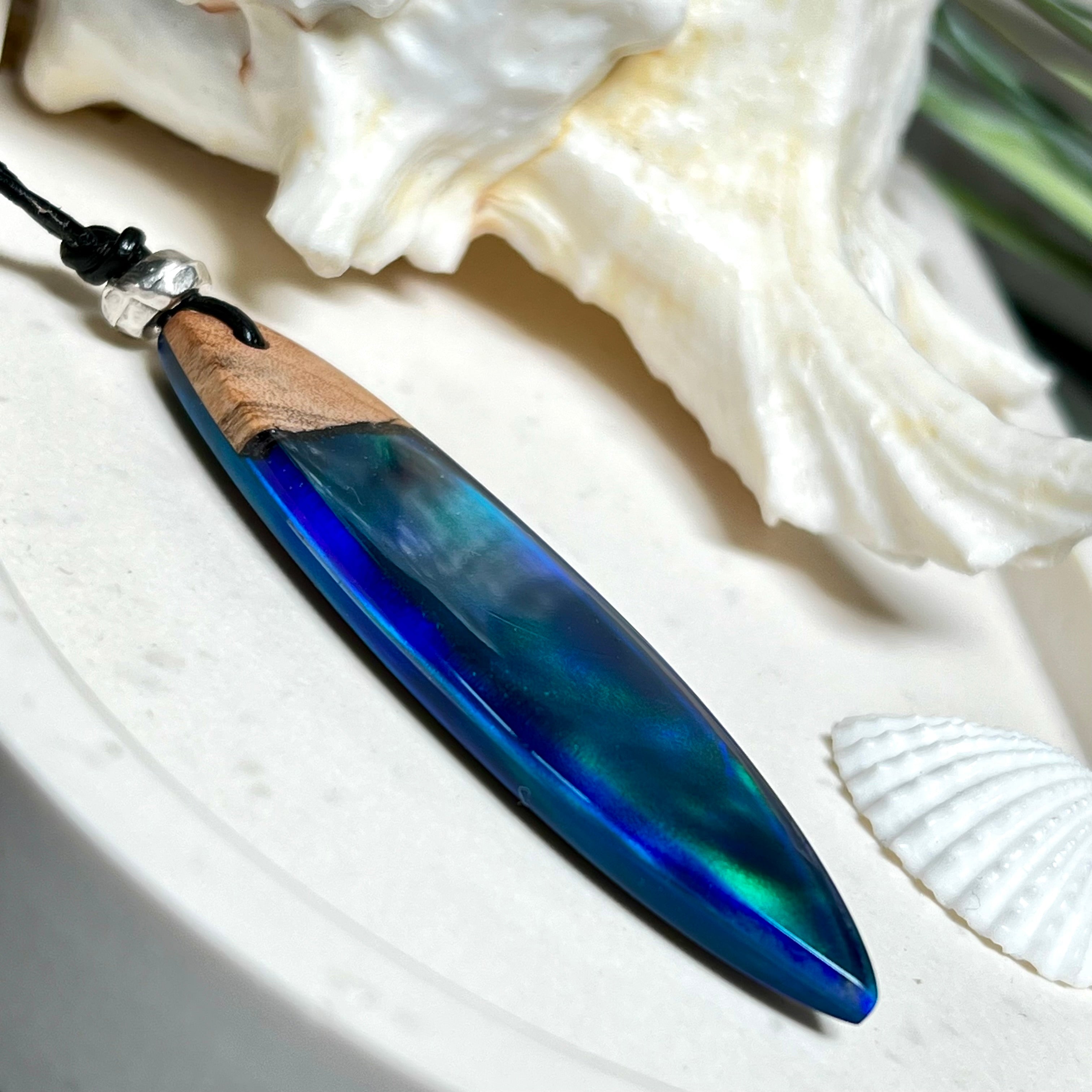 Beidseitig tragbarer Surfbrett-Anhänger aus Olivenholz, kombiniert mit klaren Epoxidharz in Blau-Grün. Eingearbeiteter synthetischer Opal erzeugt ein faszinierendes Farbspiel wie Nordlichter. Größe ca. 6 × 1,8 cm. Mit verstellbarem Lederband (26–45 cm), dekorativen Perlen & Schiebeknoten. Handgefertigt & einzigartig. Ein Schmuckstück mit Naturinspiration.
