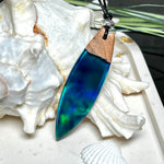Beidseitig tragbarer Surfbrett-Anhänger aus Olivenholz, kombiniert mit klaren Epoxidharz in Blau-Grün. Eingearbeiteter synthetischer Opal erzeugt ein faszinierendes Farbspiel wie Nordlichter. Größe ca. 6 × 1,8 cm. Mit verstellbarem Lederband (26–45 cm), dekorativen Perlen & Schiebeknoten. Handgefertigt & einzigartig. Ein Schmuckstück mit Naturinspiration.
