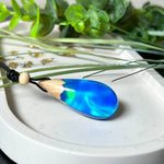 Handgefertigter Tropfenanhänger aus Maserholz und Harz mit synthetischem Opal. Die Farben schimmern je nach Lichteinfall in leuchtendem Blau und Grün – inspiriert von Polarlichtern. Der Anhänger ist ca. 3,9 × 1,8 cm groß und hängt an einem verstellbaren Lederband mit Holzperlen. Jedes Stück ist ein Unikat mit einzigartiger Holzmaserung. Ideal als Geschenk oder stilvoller Begleiter im Alltag.