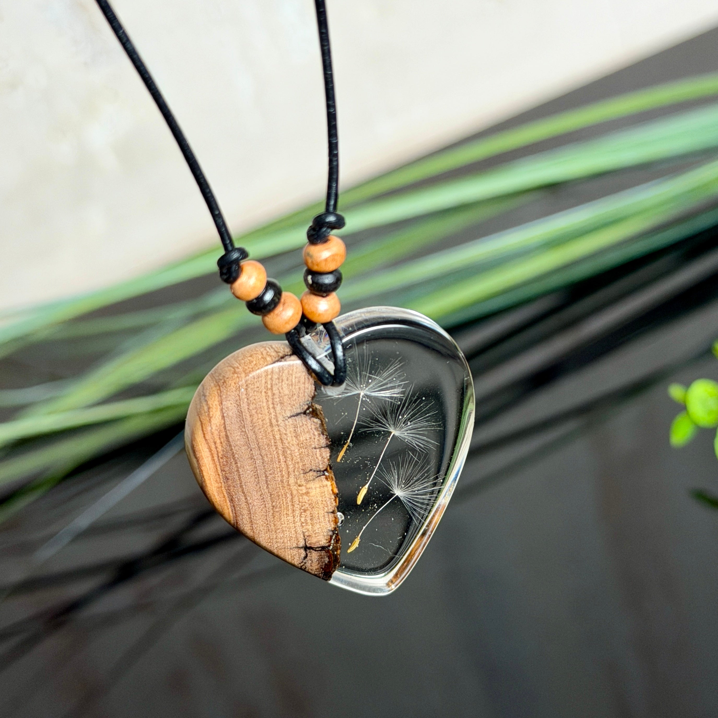 Handgefertigter Herzanhänger aus warm gemasertem Olivenholz und kristallklarem Epoxidharz mit schwebenden Pusteblumensamen. Schwarzes, verstellbares Lederband mit Schiebeknoten und naturfarbenen Holzperlen, Anhänger ca. 3,5 × 3,3 cm. Naturnaher Unikatschmuck von ZeitlosSchmuckDesign.