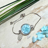 Verstellbares Edelstahlarmband mit Zirkoniasteinchen und Schiebeverschluss und rundem Anhänger (ca. 2,5 × 1,7 cm) mit echten Larimar-Edelstein-Splittern in Harz. Zusätzlich ein Charm „Blume des Lebens“ aus 925 Silber. Handgefertigtes Unikat in Türkis- und Meeresblautönen – Schmuckstück in Nahaufnahme bzw. als Tragebild am Handgelenk.