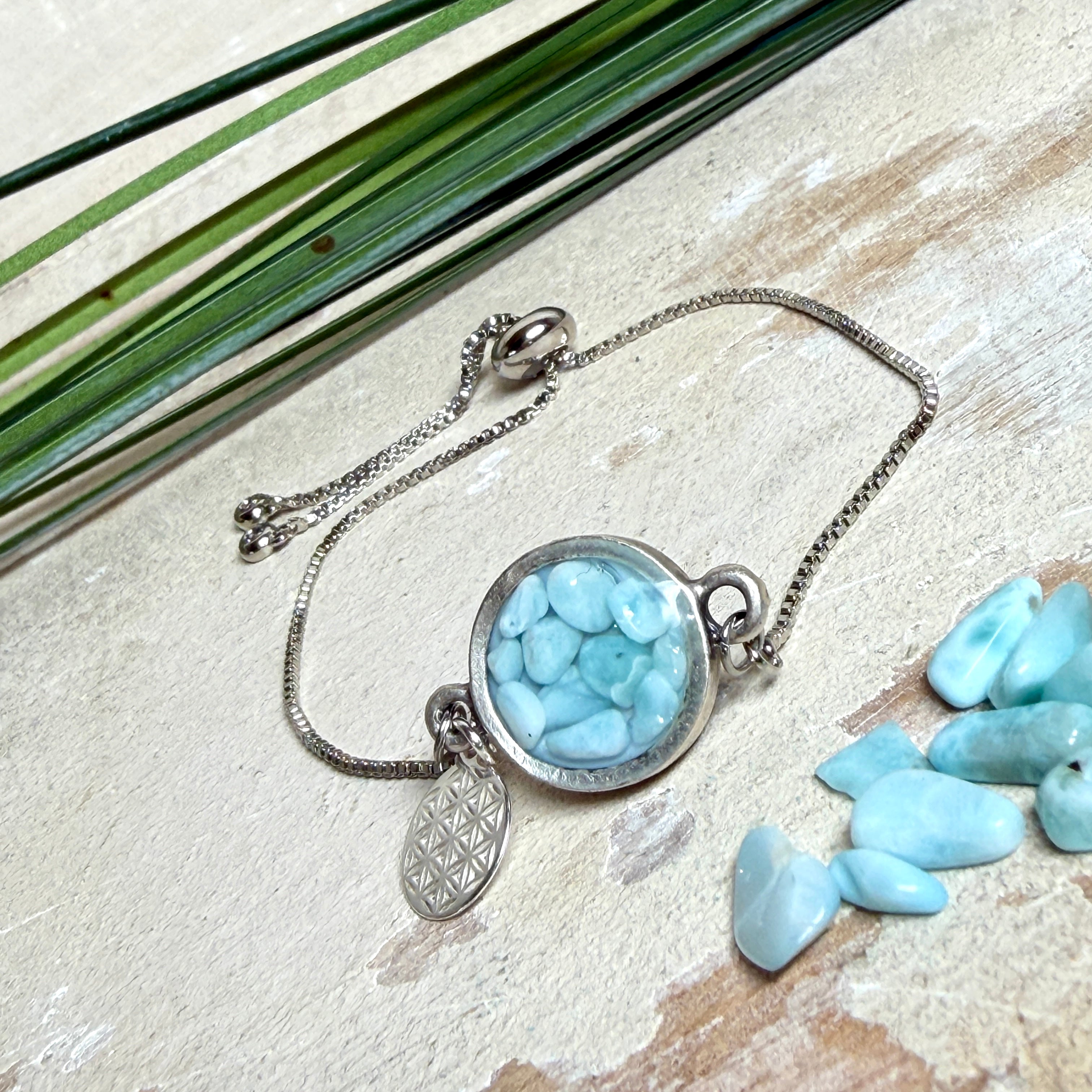 Verstellbares Edelstahlarmband mit Zirkoniasteinchen und Schiebeverschluss und rundem Anhänger (ca. 2,5 × 1,7 cm) mit echten Larimar-Edelstein-Splittern in Harz. Zusätzlich ein Charm „Blume des Lebens“ aus 925 Silber. Handgefertigtes Unikat in Türkis- und Meeresblautönen – Schmuckstück in Nahaufnahme bzw. als Tragebild am Handgelenk.