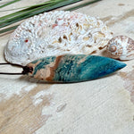 Unikat Anhänger aus stabilisiertem Holz mit türkisblauem Harzverlauf, schlanke Form, poliert. Verstellbares braunes Lederband 22–46 cm mit Schiebeknoten. Anhänger ca. 6,6 × 2 cm. Tragefoto am Hals und Produktfotos mit maritimer Deko