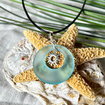 Handgefertigte Halskette mit Donut-Anhänger aus türkisfarbenem Seeglas, kombiniert mit einem filigranen Sonnen-Charm aus Edelstahl. Die Kette besteht aus einer schwarzen Lederschnur (1,5 mm) mit Karabiner und Verlängerungskette aus Edelstahl. Maritimes Schmuckstück für Meerliebhaberinnen und Sommerfreunde. Unikat mit stilvoller Ausstrahlung.
