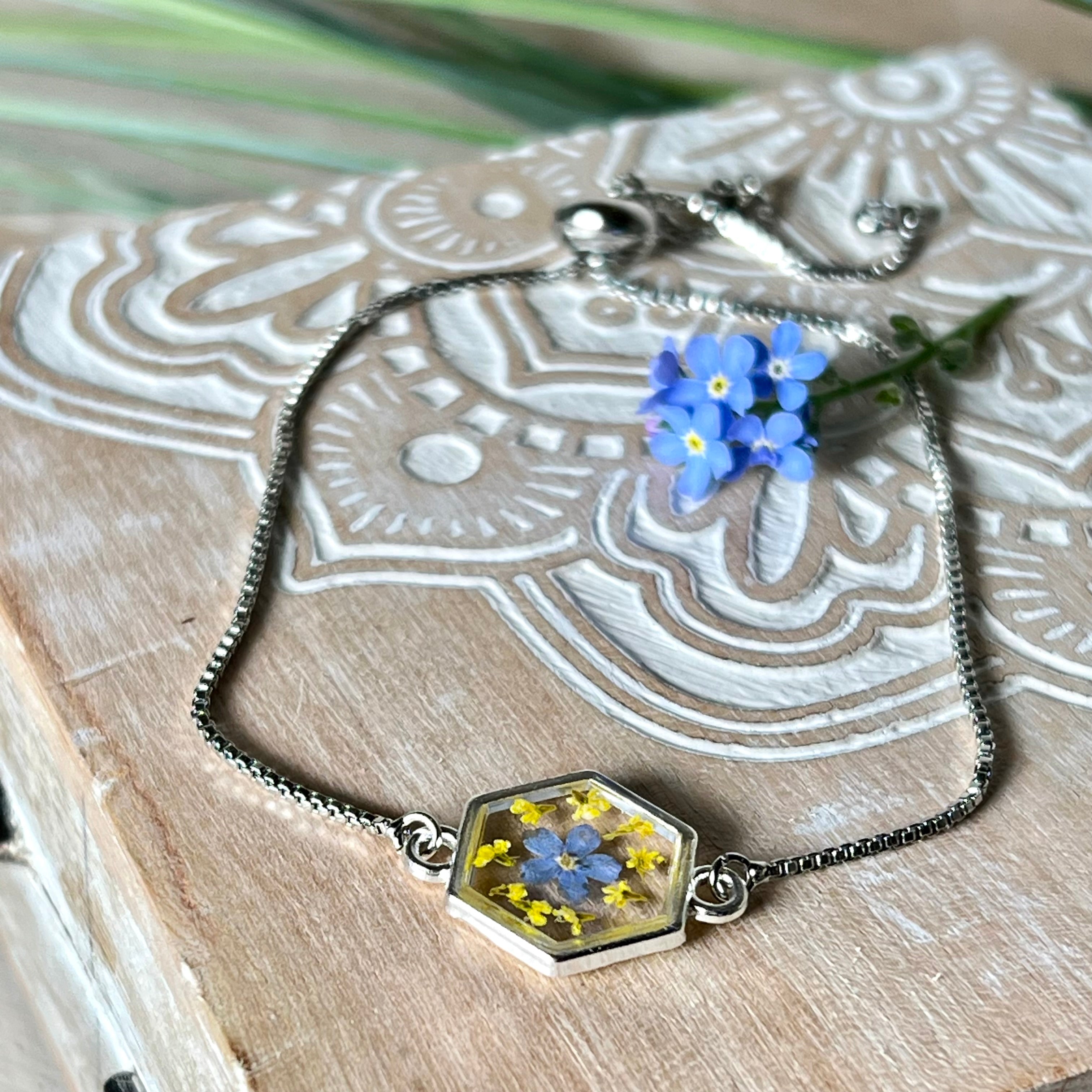 „Detailaufnahme eines handgefertigten Armbands mit echter blauer Vergissmeinnicht und feinen gelben Blüten in Harz, eingefasst in sechseckiger versilberter Lünette. Verstellbares Edelstahlarmband mit funkelnden Zirkonia-Endperlen – liebevolles Natur-Schmuckstück, ideal als Geschenk für Frauen.“