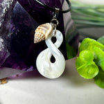 Detailaufnahme eines handgefertigten Anhängers aus echtem Perlmutt in Schlaufenform, kombiniert mit einer kleinen echten Muschel als Charm-Anhänger. Der Schmuck hängt an einer feinen schwarzen Lederschnur mit Edelstahlverschluss und symbolisiert Harmonie und Unendlichkeit. Ideal für Meeresliebhaberinnen und Fans von Naturmaterialien.