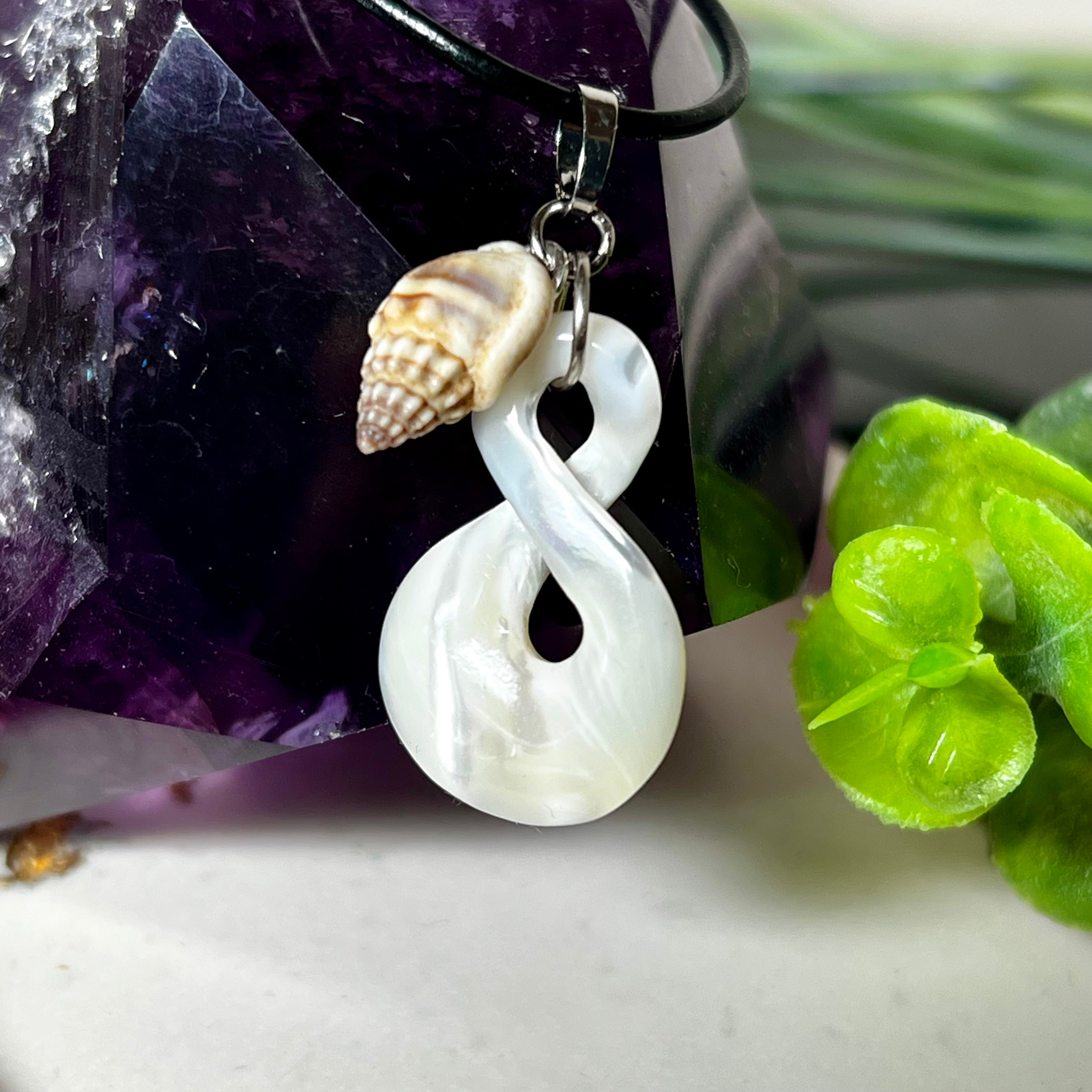 Detailaufnahme eines handgefertigten Anhängers aus echtem Perlmutt in Schlaufenform, kombiniert mit einer kleinen echten Muschel als Charm-Anhänger. Der Schmuck hängt an einer feinen schwarzen Lederschnur mit Edelstahlverschluss und symbolisiert Harmonie und Unendlichkeit. Ideal für Meeresliebhaberinnen und Fans von Naturmaterialien.