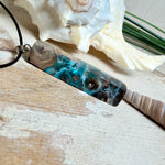 Rechteckiger Stabanhänger aus Maserholz und glasklarem Epoxidharz in tiefen Blau- und Türkistönen mit organischer Lagunen/Riff-Optik. Hochglanzpoliert, ca. 5,3 × 1,2 cm. Der Anhänger hängt an einer schwarzen Lederkette mit Edelstahl-Karabiner und 5-cm-Verlängerung (45 + 5 cm); die Anhängerschlaufe ist ebenfalls aus Edelstahl. Handgefertigtes Unikat, Lieferung in edler Schmuckbox – maritimer Look.