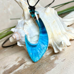 Handgefertigter Haken-Anhänger im Hei-Matau-Stil aus Maserholz und klarem Harz mit Blau-, Türkis- und Weißverlauf, hochglanzpoliert. Oben sichtbares Holz im Harzbereich, getragen an verstellbarem schwarzem Lederband mit Schiebeknoten und Zierperlen. Anhänger ca. 7 × 3 cm, fotografiert mit Muscheln im Hintergrund.