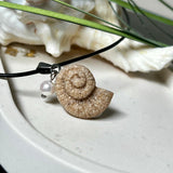 Handgefertigter Anhänger in Ammonit-Form aus echtem Meersand und Harz, kombiniert mit einer weißen Süßwasserperle. Der maritime Anhänger hängt an einer schwarzen Lederkette mit Edelstahlöse und Karabinerverschluss. Im Hintergrund liegt eine weiße Muschel, die das natürliche, strandinspirierte Design des Schmuckstücks unterstreicht. Ideal als besonderes Geschenk oder Erinnerung an den Sommer.