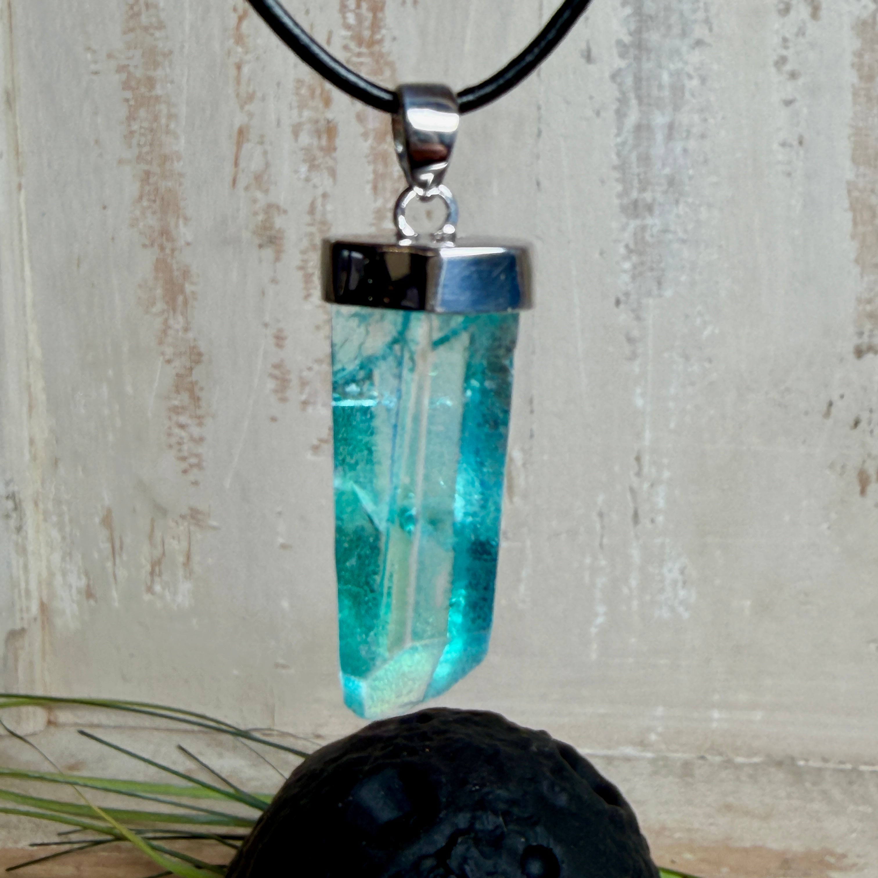 Nahaufnahme einer handgefertigten Halskette mit Aqua Aura Rohkristall: naturbelassener, sechskantiger Bergkristall mit goldbedampfter Aqua-Beschichtung in Türkisblau, gefasst in 925er Silber und getragen an einer schwarzen Lederkette mit Edelstahl-Karabiner und Verlängerungskettchen.