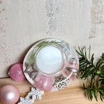 Runder Teelichthalter aus klarem Epoxidharz, Durchmesser ca. 7 cm, Höhe ca. 4 cm. Im Harz sind rosa Weihnachtskugeln, ein Stern, eine Schneeflocke, ein Tannenbaum, künstliche Tannennadeln und feiner Glitzer eingegossen. Vertiefung für ein Teelicht in der Mitte, Unterseite mit vier Korkpads als Möbelschoner. Fotografiert auf Holz mit Tannenzweig und rosa Kugeln als weihnachtliche Dekoration