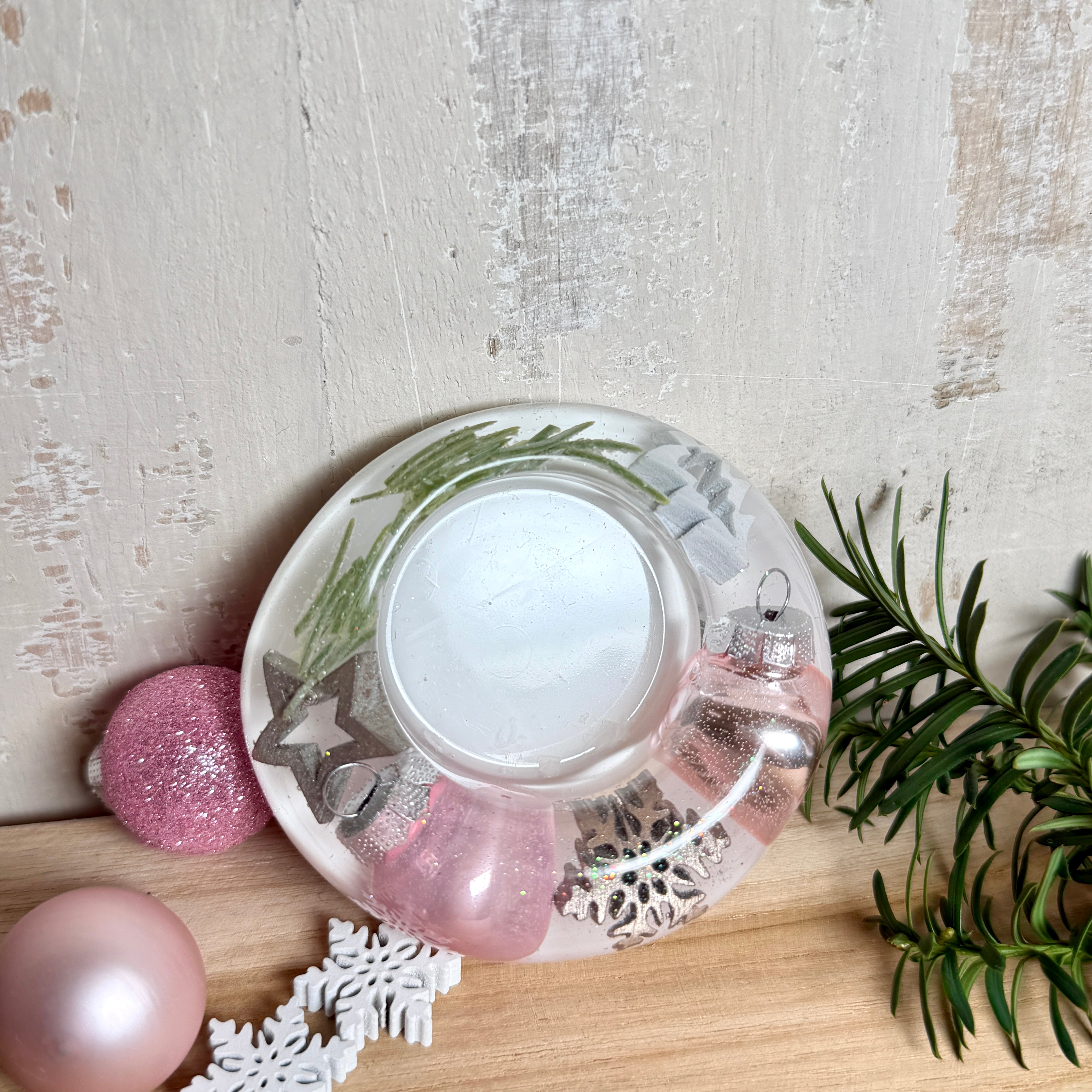 Runder Teelichthalter aus klarem Epoxidharz, Durchmesser ca. 7 cm, Höhe ca. 4 cm. Im Harz sind rosa Weihnachtskugeln, ein Stern, eine Schneeflocke, ein Tannenbaum, künstliche Tannennadeln und feiner Glitzer eingegossen. Vertiefung für ein Teelicht in der Mitte, Unterseite mit vier Korkpads als Möbelschoner. Fotografiert auf Holz mit Tannenzweig und rosa Kugeln als weihnachtliche Dekoration