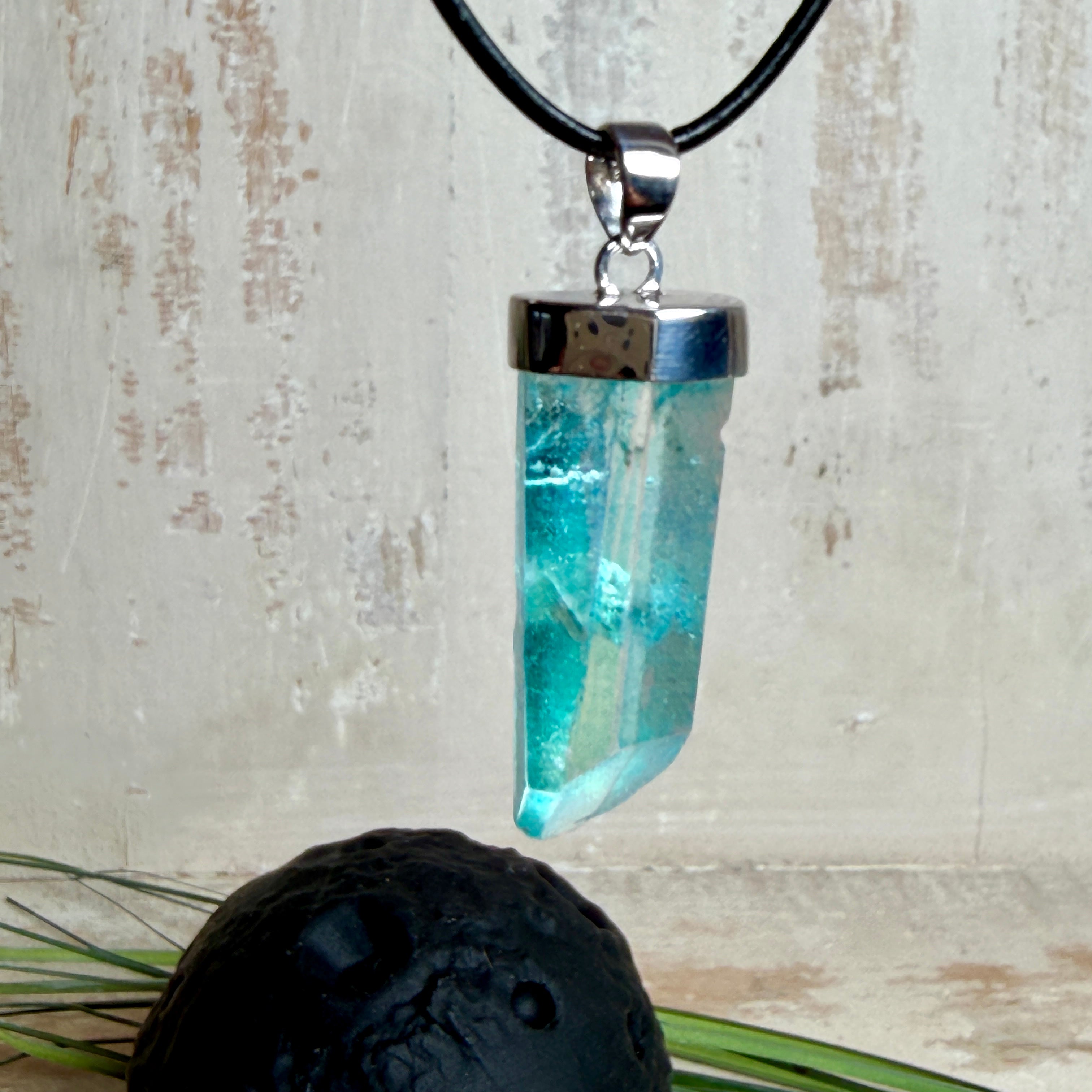 Nahaufnahme einer handgefertigten Halskette mit Aqua Aura Rohkristall: naturbelassener, sechskantiger Bergkristall mit goldbedampfter Aqua-Beschichtung in Türkisblau, gefasst in 925er Silber und getragen an einer schwarzen Lederkette mit Edelstahl-Karabiner und Verlängerungskettchen.