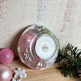 Runder Teelichthalter aus klarem Epoxidharz, Durchmesser ca. 7 cm, Höhe ca. 4 cm. Im Harz sind rosa Weihnachtskugeln, ein Stern, eine Schneeflocke, ein Tannenbaum, künstliche Tannennadeln und feiner Glitzer eingegossen. Vertiefung für ein Teelicht in der Mitte, Unterseite mit vier Korkpads als Möbelschoner. Fotografiert auf Holz mit Tannenzweig und rosa Kugeln als weihnachtliche Dekoration