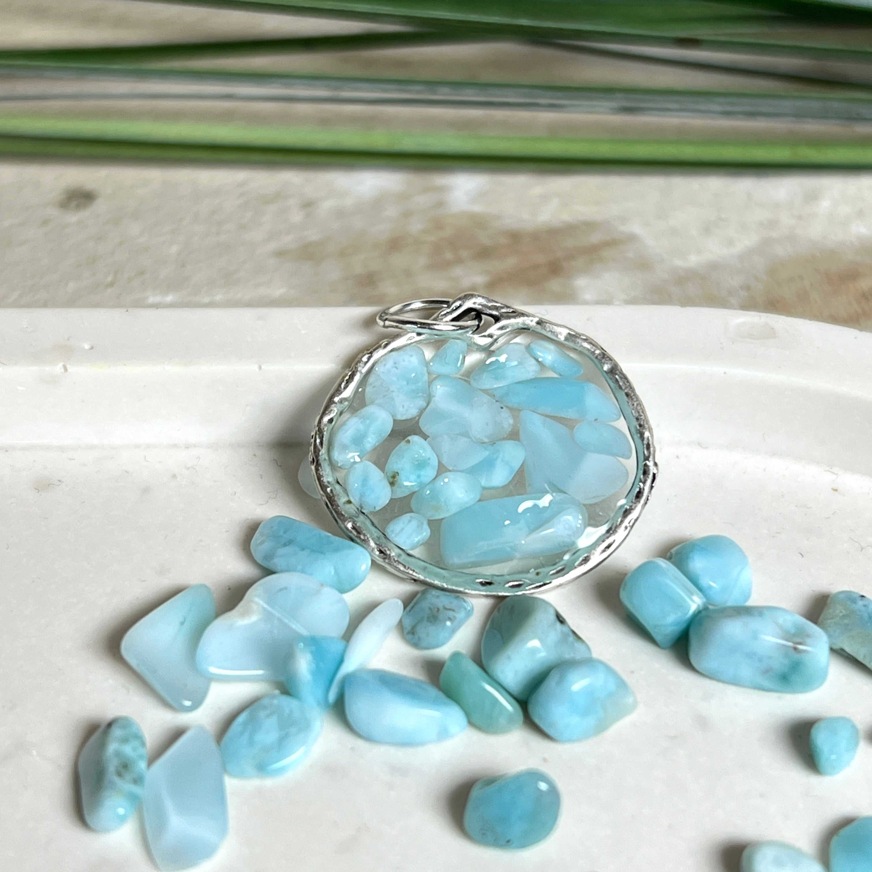 Runder Anhänger „Atlantis Dream“ aus antik-silberfarbener Zinklegierung, gefüllt mit kleinen Larimar-Edelsteinchen und klarem Resin. Die Larimarsteine zeigen sanfte Blau- und Weißtöne, die an das Meer erinnern. Durchmesser ca. 22 mm, handgefertigtes Unikat, fotografiert auf maritimem Untergrund mit kleinen Larimarsteinen und Muscheln zur Größenorientierung.