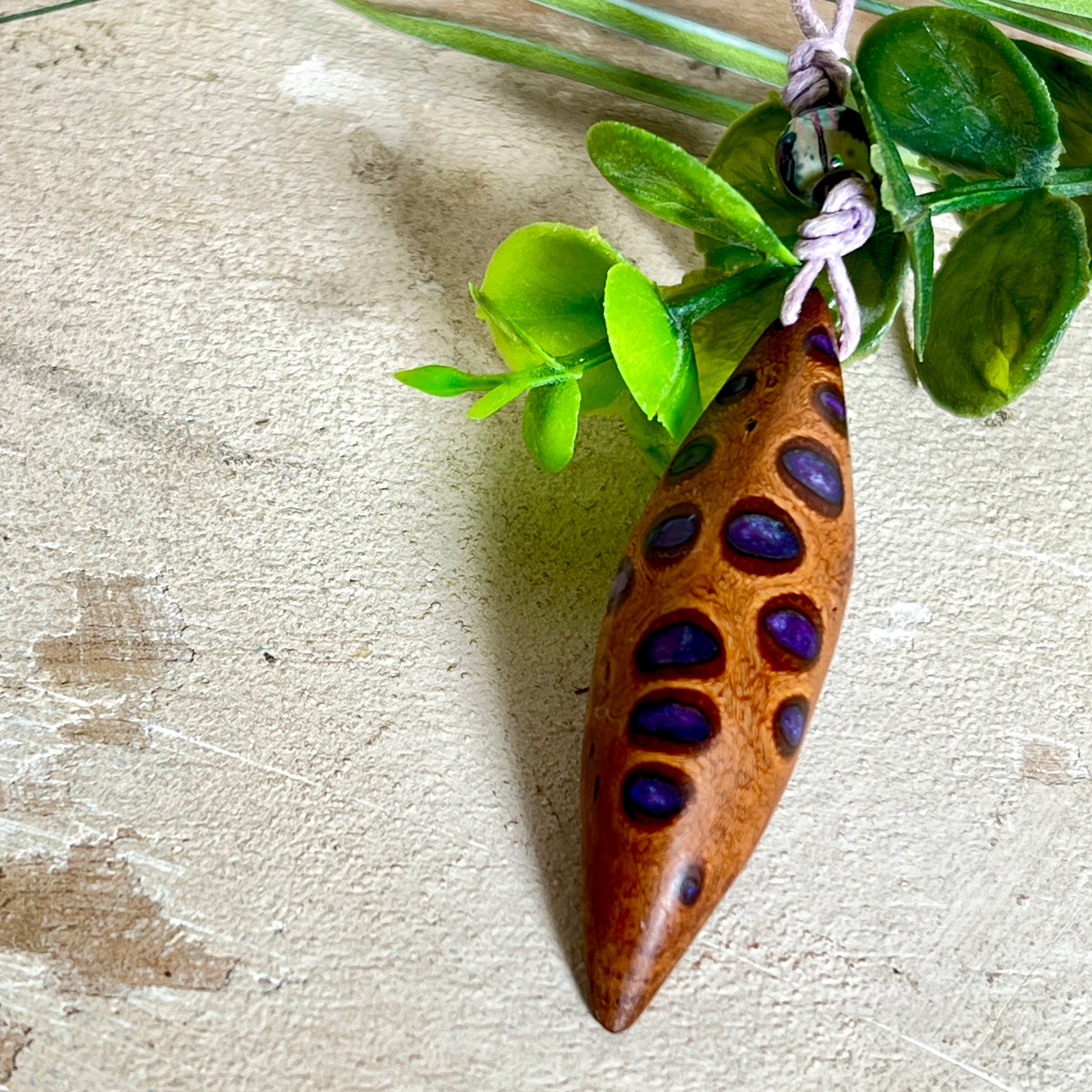 Handgefertigter Anhänger aus einem australischen Banksiazapfen mit eingelassenem metallic-violettem Harz. Der ovale, tropfenförmige Anhänger ist ca. 5,5 cm lang und wird mit einem verstellbaren Baumwollband geliefert. Jedes Stück zeigt die einzigartige natürliche Struktur des Zapfens und schimmert in satten Lilatönen – ein stilvoller Schmuck mit organischem Charakter.
