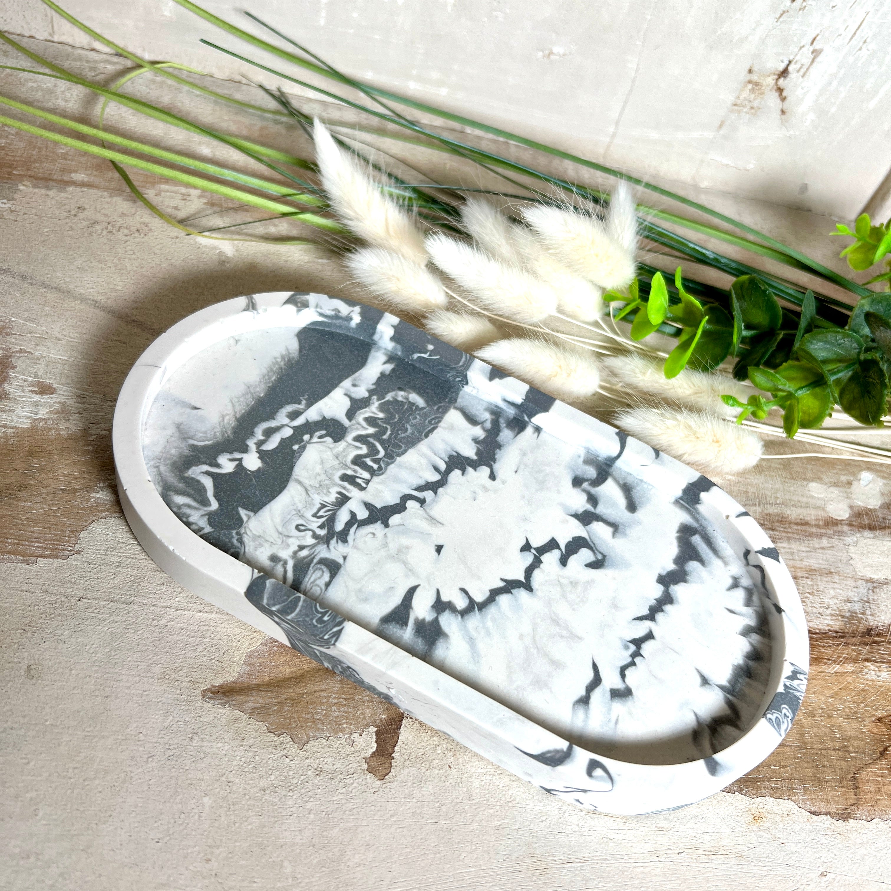 Handgegossenes Schmucktablett aus Jesmonite mit grau -weißer Marmorierung. Die ovale Form und die individuelle Musterung machen jedes Stück einzigartig. Die Rückseite ist mit rutschfesten Korkpunkten ausgestattet – ideal als stilvolle Ablage für Schmuck, Kristalle, Schlüssel oder Brille. Unikat in modernem Monochrom-Design. Lieferung ohne Deko.