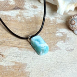 Larimar Freeform Anhänger an schwarzer Lederkette. Natürlicher, rechteckiger Larimar-Stein (ca. 1,5 × 1 cm) in Türkis-Blau mit weißen Strukturen, eingefasst in eine rhodinierte Silberöse. Beidseitig tragbar, an 45 cm Lederkette mit Edelstahl-Karabinerverschluss und 5 cm Verlängerung. Maritimes, zurückhaltend elegantes Schmuckstück mit der Optik karibischen Meerwassers.