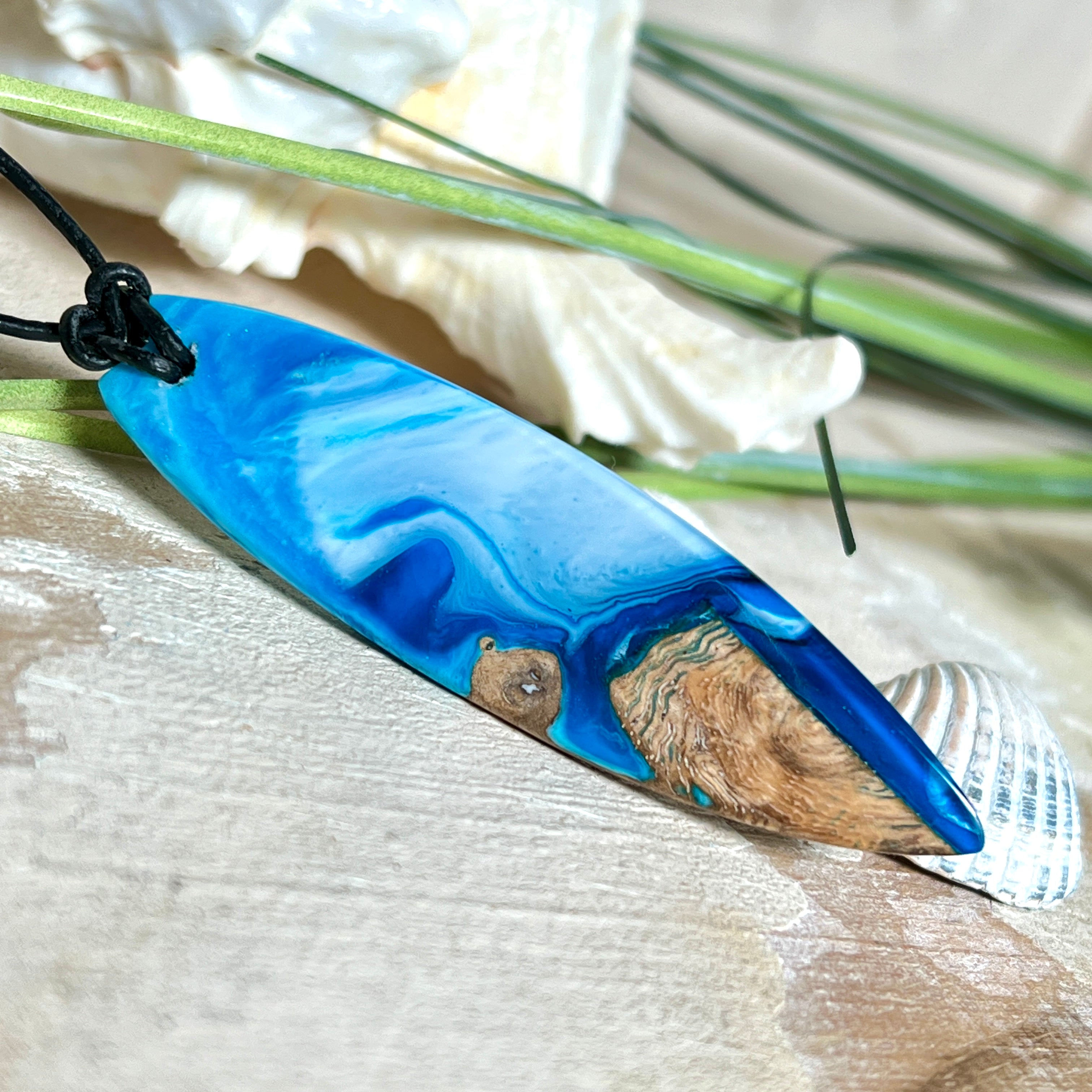 Handgefertigter Surfbrett-Anhänger „Ocean Glide“ aus Maserholz und Epoxidharz in verschiedenen Blau-, und Weißtönen. Der ca. 6,5 × 2 cm große Anhänger wird an einem verstellbaren Lederband (26–46 cm) mit Holzperlen geliefert. Ein maritimes Unikat mit natürlicher Holzmaserung, das die Schönheit des Meeres einfängt.