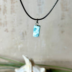 Larimar Freeform Anhänger an schwarzer Lederkette. Natürlicher, rechteckiger Larimar-Stein (ca. 1,5 × 1 cm) in Türkis-Blau mit weißen Strukturen, eingefasst in eine rhodinierte Silberöse. Beidseitig tragbar, an 45 cm Lederkette mit Edelstahl-Karabinerverschluss und 5 cm Verlängerung. Maritimes, zurückhaltend elegantes Schmuckstück mit der Optik karibischen Meerwassers.