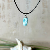 Larimar Freeform Anhänger an schwarzer Lederkette. Natürlicher, rechteckiger Larimar-Stein (ca. 1,5 × 1 cm) in Türkis-Blau mit weißen Strukturen, eingefasst in eine rhodinierte Silberöse. Beidseitig tragbar, an 45 cm Lederkette mit Edelstahl-Karabinerverschluss und 5 cm Verlängerung. Maritimes, zurückhaltend elegantes Schmuckstück mit der Optik karibischen Meerwassers.