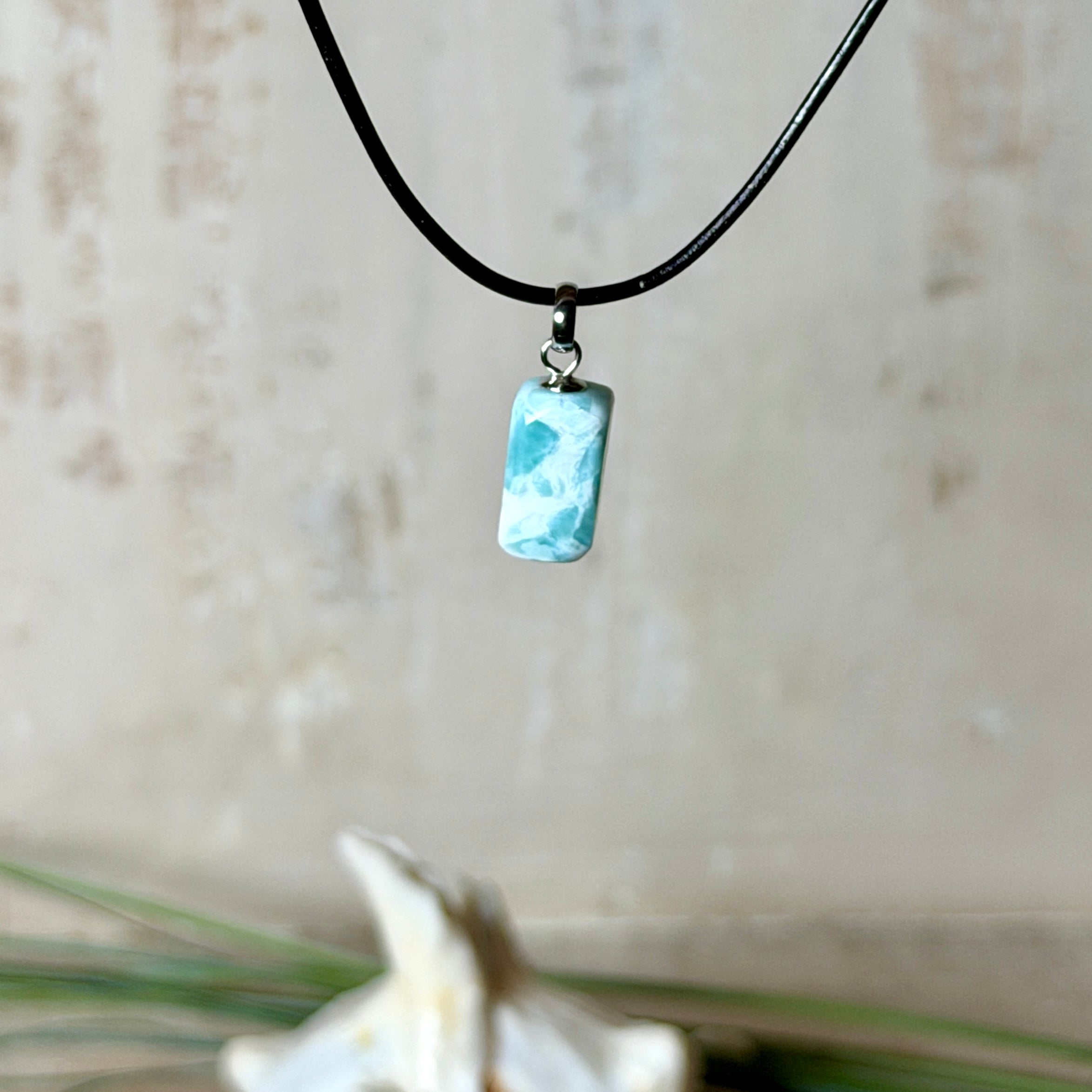 Larimar Freeform Anhänger an schwarzer Lederkette. Natürlicher, rechteckiger Larimar-Stein (ca. 1,5 × 1 cm) in Türkis-Blau mit weißen Strukturen, eingefasst in eine rhodinierte Silberöse. Beidseitig tragbar, an 45 cm Lederkette mit Edelstahl-Karabinerverschluss und 5 cm Verlängerung. Maritimes, zurückhaltend elegantes Schmuckstück mit der Optik karibischen Meerwassers.