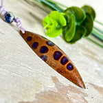 Handgefertigter Anhänger aus einem australischen Banksiazapfen mit eingelassenem metallic-violettem Harz. Der ovale, tropfenförmige Anhänger ist ca. 5,5 cm lang und wird mit einem verstellbaren Baumwollband geliefert. Jedes Stück zeigt die einzigartige natürliche Struktur des Zapfens und schimmert in satten Lilatönen – ein stilvoller Schmuck mit organischem Charakter.