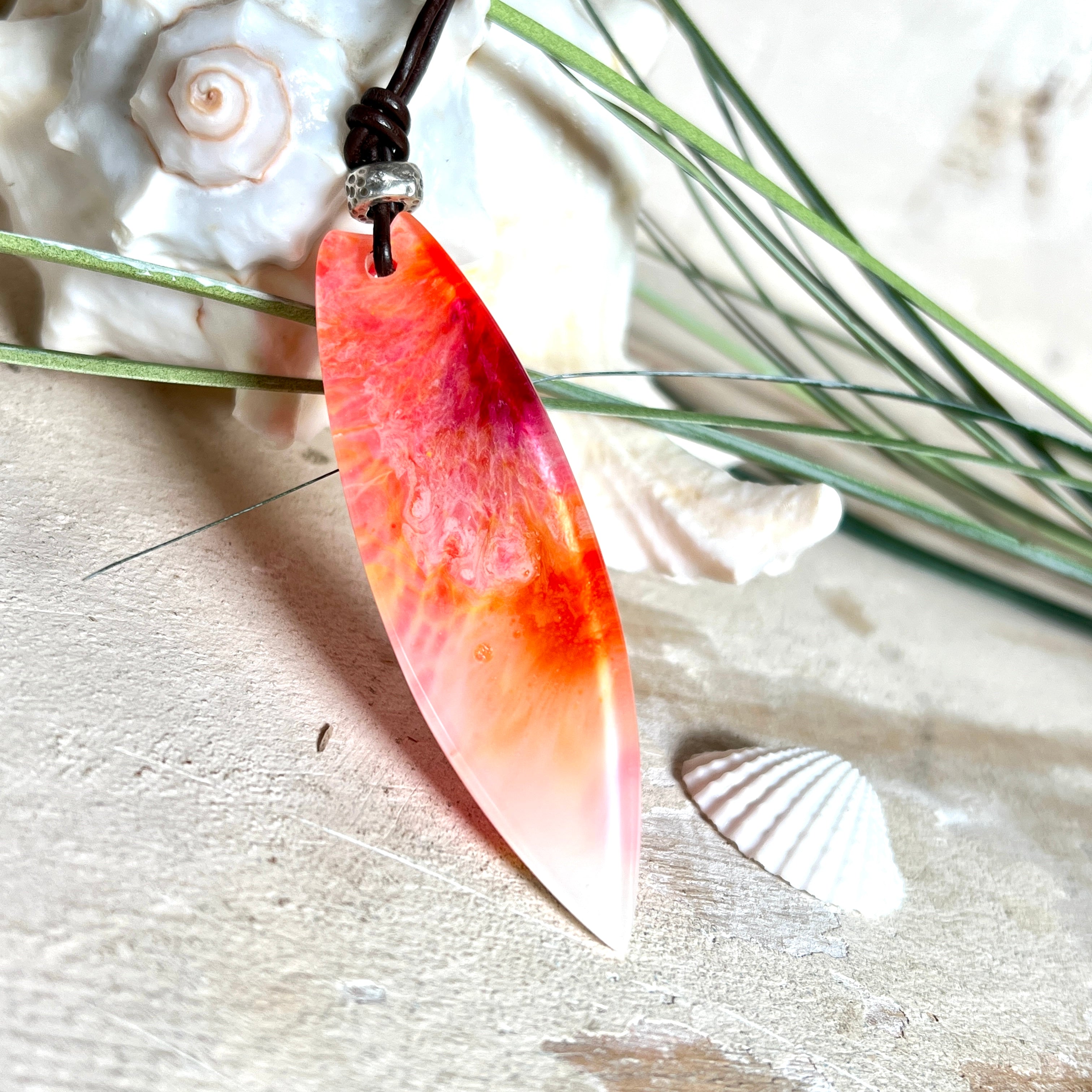 Handgefertigter Surfbrett-Anhänger aus fluoreszierendem Epoxidharz in leuchtenden Orange-, Rot- und Gelbtönen. Leuchtet bei Schwarzlicht. Kombiniert mit verstellbarem Lederband (26–45 cm), verziert mit Spacer- und Holzperlen. Das Design ist inspiriert von der Farbintensität eines Sonnenuntergangs über dem Meer.