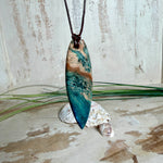 Unikat Anhänger aus stabilisiertem Holz mit türkisblauem Harzverlauf, schlanke Form, poliert. Verstellbares braunes Lederband 22–46 cm mit Schiebeknoten. Anhänger ca. 6,6 × 2 cm. Tragefoto am Hals und Produktfotos mit maritimer Deko