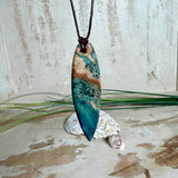 Unikat Anhänger aus stabilisiertem Holz mit türkisblauem Harzverlauf, schlanke Form, poliert. Verstellbares braunes Lederband 22–46 cm mit Schiebeknoten. Anhänger ca. 6,6 × 2 cm. Tragefoto am Hals und Produktfotos mit maritimer Deko