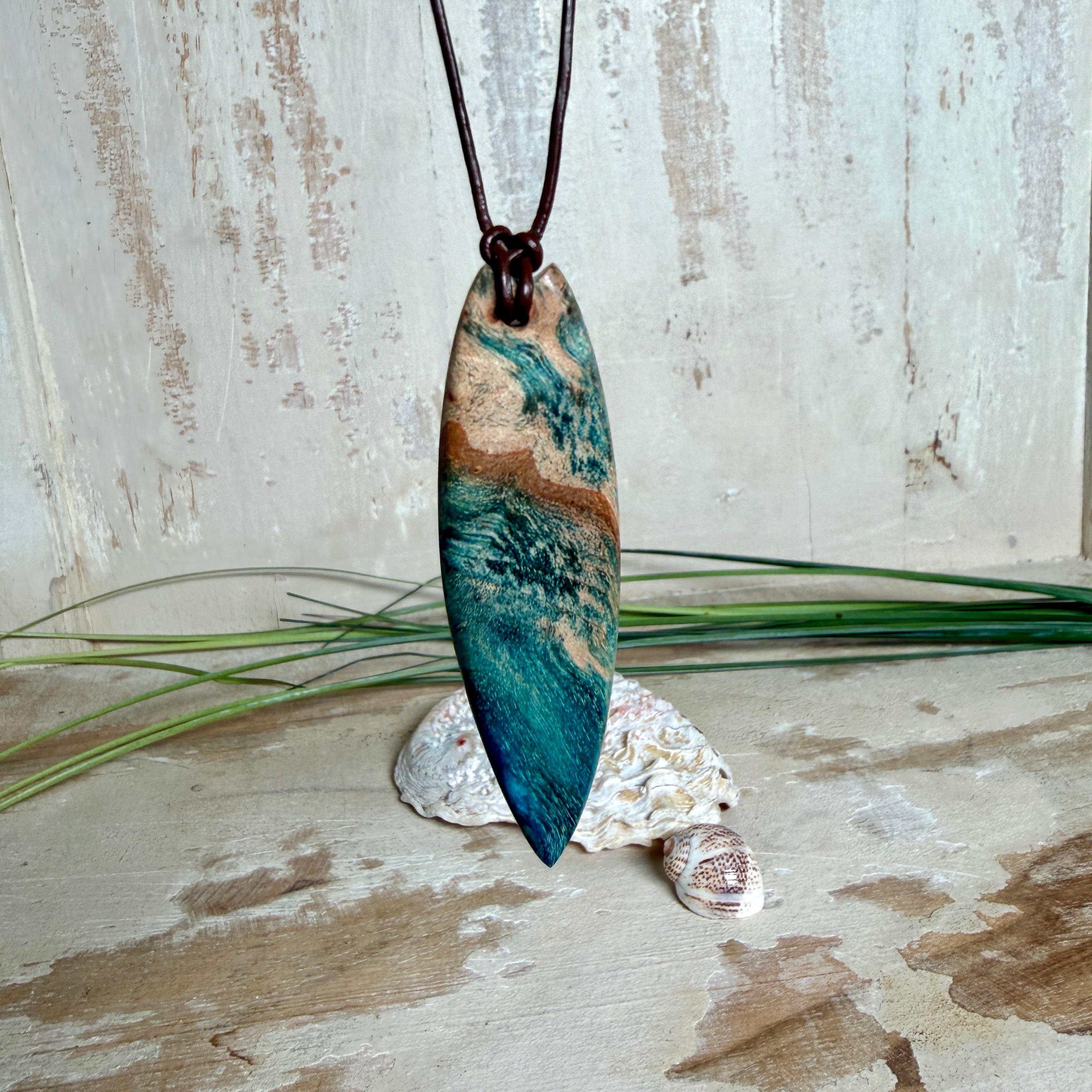 Unikat Anhänger aus stabilisiertem Holz mit türkisblauem Harzverlauf, schlanke Form, poliert. Verstellbares braunes Lederband 22–46 cm mit Schiebeknoten. Anhänger ca. 6,6 × 2 cm. Tragefoto am Hals und Produktfotos mit maritimer Deko