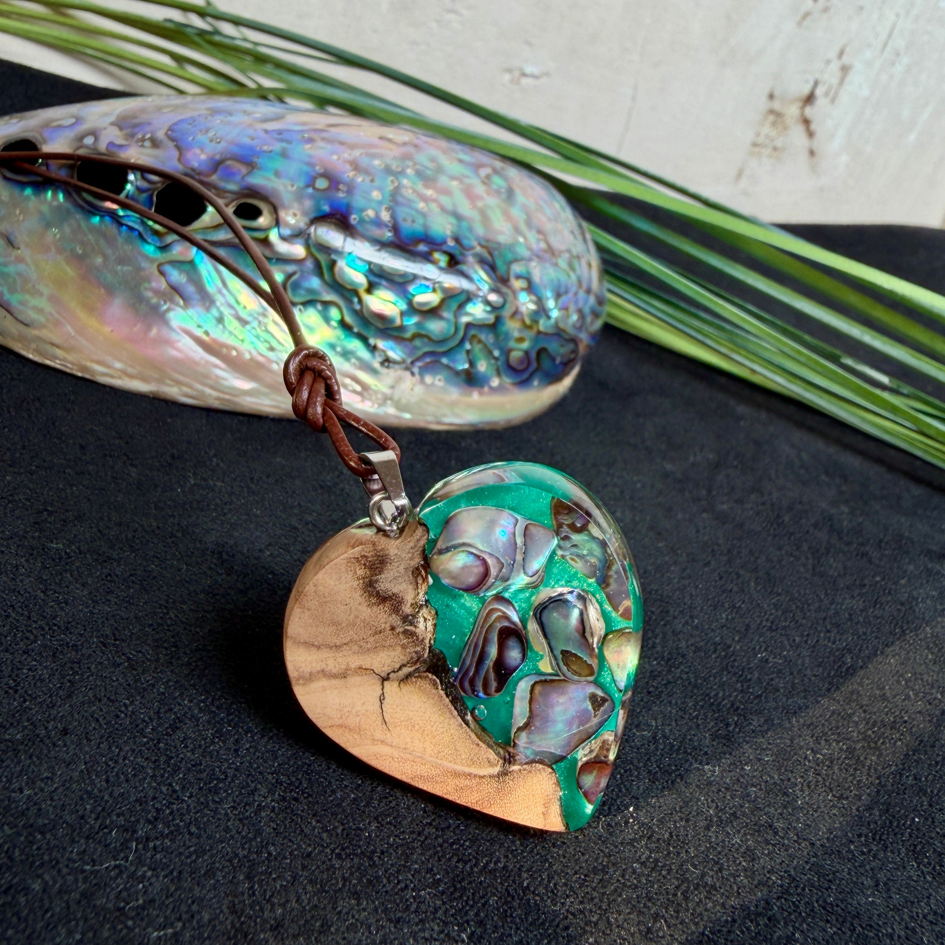 Herzförmiger Anhänger aus Olivenholz mit aquamarinfarbenem Harz und eingelegten Paua-Muschelstücken (Abalone), irisierender Blau-Grün-Schimmer. Der Anhänger hängt an einem braunen, verstellbaren Lederband mit Schiebeknoten; Aufhängung aus Edelstahl. Produktfoto vor maritimer Deko/Abalone-Schale.